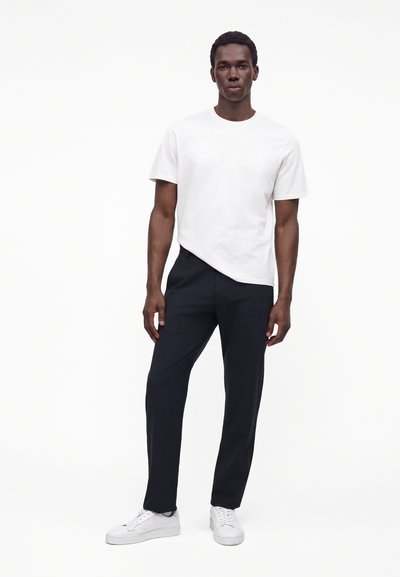 Homme debout portant un t-shirt blanc uni à manches courtes, un pantalon noir et des baskets blanches sur fond blanc.