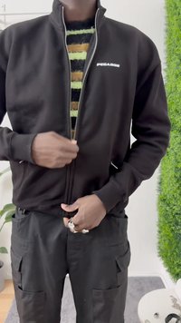 Giacca nera con zip e logo, presenta un colletto alto, tasche laterali e polsini a costine. Indossata sopra un maglione a righe verde e marrone.