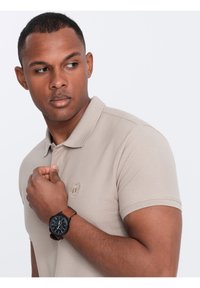 Beige polokošile s krátkým rukávem, texturovaná tkanina a vyšívané logo. Černé hodinky s hnědým koženým páskem na zápěstí.