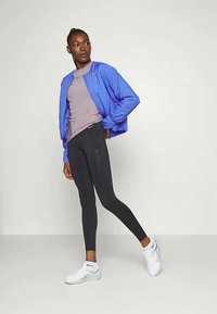 Jaqueta bomber leve azul sobre uma blusa lilás clara, combinada com leggings pretas e tênis atléticos brancos. Tecido suave, design ajustado.