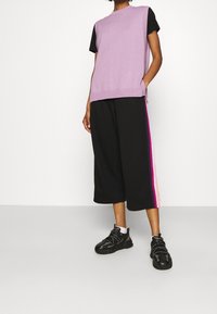 Fioletowy dzianinowy top na ramiączkach, nałożony na czarną koszulę z krótkim rękawem, w zestawieniu z czarnymi spodniami o szerokich nogawkach z kolorową paskiem. Czarne sneakersy.