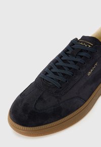 Tenis navy din suede cu un exterior texturat, cusături albastre și o talpă din cauciuc de tip gumă. Dispune de un sistem de închidere cu șireturi și de un logo auriu pe limbă.