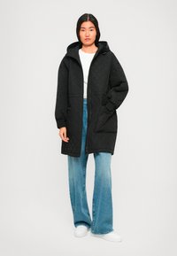 Jongeman staat in een gewatteerde zwarte hoodiejas, witte blouse, wijde blauwe jeans en witte sneakers tegen een effen achtergrond.