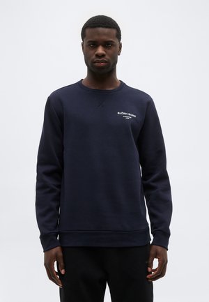 Ung mand iført en marineblå Björn Borg-sweatshirt og sorte bukser, stående mod en ensartet lysegrå baggrund.