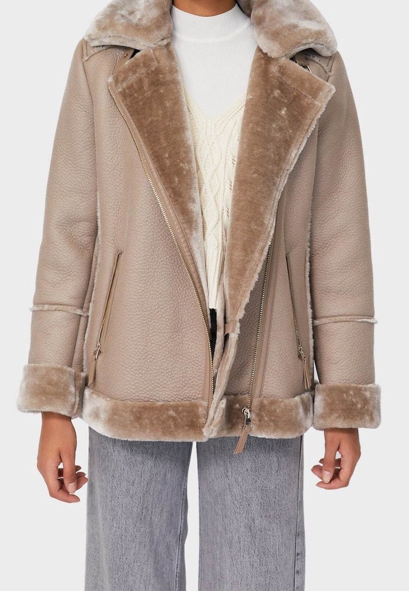 Veste en shearling beige avec col, poignets et ourlet en peluche, portée sur un pull en maille blanc et un jean gris.