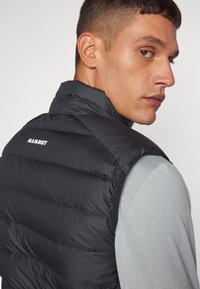 Zwarte puffer vest met horizontale stiksels, hoge kraag en Mammut-logo op de rug. Draagt over een lichtgrijs shirt, strakke ontwerp.
