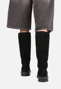 Black suede knee-high boots with a smooth texture, side seams, and thick rubber soles. The boots contrast with gray high-waisted shorts.