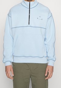 Světle modrý svetr s půlzipem, černým výrazným švem, klokaní kapsou a malým logem na hrudi.