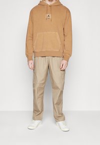 Felpa marrone con cordino, tasca frontale e logo; abbinata a pantaloni cargo beige; scarpe da ginnastica bianche con suole testurizzate.