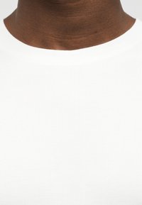 Primo piano del collo e della parte superiore del torace di una persona che indossa una maglietta bianca semplice a costine con scollo tondo.