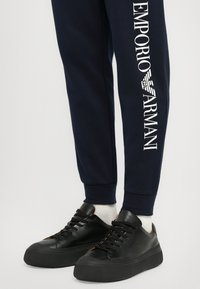Navy joggers with white "Emporio Armani" text on the left leg, paired with black sneakers featuring thick soles and orange accents.