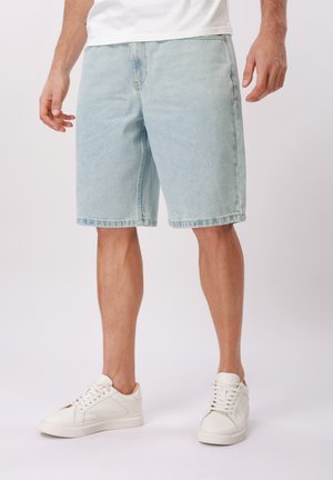 Mand iført lyseblå denimshorts, hvide sneakers og hvid skjorte, står foran en ensfarvet hvid baggrund.