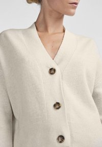 Μπεζ πλεκτό κασκόλ με V-λαιμό, τρία σκούρα κουμπιά και ριχτή υφή. Απαλή ύφανση με άνετη εφαρμογή.