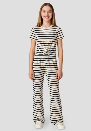 Fille souriante debout, vêtue d’un t-shirt et pantalon assortis crème et noir à rayures horizontales, avec des baskets blanches sur fond gris uni.