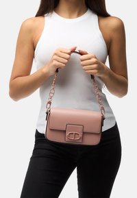 Jasnoróżowa skórzana torebka o prostokątnym kształcie, z przednią klapą, metalowym logo i łańcuszkowym paskiem. Gładka faktura i minimalistyczny design.