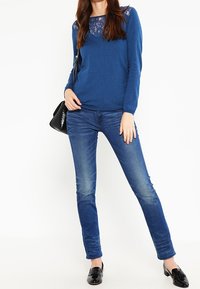 Sininen pitkähihainen neulepaita, jossa on pitsikoristelu pääntiessä, yhdistetty kapealahkeisiin sinisiin farkkuihin ja mustiin loafer-kenkiin. Teksturoidu kangas kauttaaltaan.