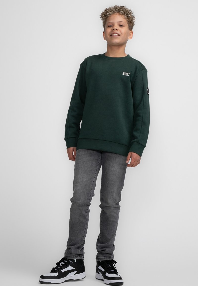 Felpa verde con scollatura rotonda, polsini a coste e un piccolo logo sul petto, abbinata a jeans grigi e sneakers nere.