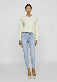 Suéter de canalé crema con cuello redondo, combinado con jeans ajustados azul claro y tacones beige de punta. Diseño simple y contemporáneo.