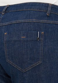 Dark blue denim jeans with a visible back pocket, contrasting orange stitching, and a small black accent detail near the pocket edge.