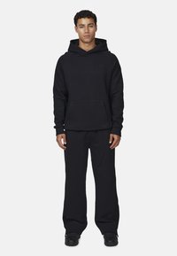 Felpa nera con cappuccio e pantaloni tuta abbinati in tessuto spesso, caratterizzati da una tasca frontale e un dettaglio logo sul petto. Vestibilità casual, design minimal.
