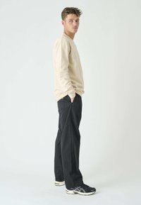 Beige long-sleeve shirt with crew neck, black tapered pants, and black athletic shoes with white accents. Model poses sideways with hands in pockets.