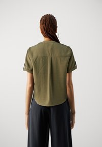 Kvinde med fletninger, iført hoop øreringe, en olivengrøn T-shirt med korte ærmer og sorte vide bukser, stående foran en ensfarvet baggrund.