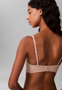 Mujer con cabello oscuro y rizado que lleva un sujetador beige claro, vista desde atrás sobre un fondo gris liso.
