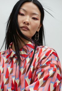Colorful, patterned shirt in red, pink, brown, and blue with a collar and long sleeves. Features a smooth texture and geometric design.