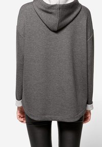 Widok z tyłu osoby ubranej w szary sweatshirt z kapturem i białymi mankietami oraz czarne dopasowane spodnie na jednolitym tle.
