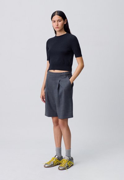 Suéter negro corto, pantalones cortos grises con pliegues y botones laterales, calcetines grises y zapatillas plateadas con cordones amarillos. Diseño simple, corte ajustado.