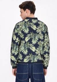 Giubbotto bomber navy con stampa a foglie di palma verdi, colletto e polsini in maglia, mostrato da dietro. Realizzato in tessuto morbido e leggero.