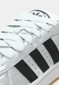 Sidabrinis sportbačis su juodomis juostelėmis, turintis plokščius šviesiai mėlynus raištelius ir blizgią tekstūrą. Yra perforuotos detalės ir logotipas ant liežuvėlio.