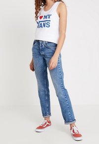 Crop top blanc sans manches avec texte bleu « I LOVE MY PANTS », associé à un jean droit bleu clair et des baskets Vans rouges.