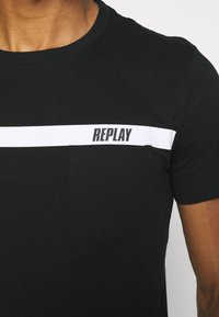 Camiseta de algodón negra con una franja horizontal blanca con "REPLAY" impreso en negro. Incluye un bolsillo parche en el lado izquierdo.