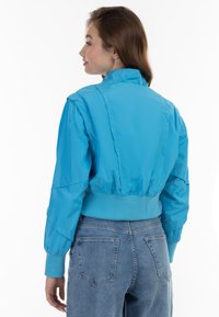 Giacca bomber blu brillante, tagliata, con colletto alto, polsini a coste e vita elastica. Tessuto liscio e lucido, design minimalista, vestibilità casual.