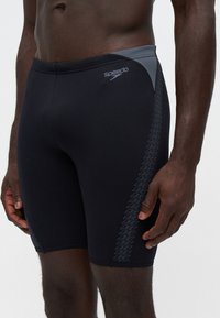 Мъж, облечен в черни и сиви плувни гащета Speedo с зигзагообразен модел отстрани, стои на светъл фон.