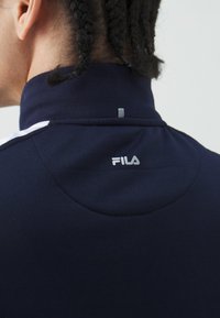 Marineblauw kledingstuk met lange mouwen en een hoge kraag, voorzien van een Fila-logo en subtiele stikdetails op de rug. Glad, rekbaar materiaal.