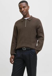 Muž v hnědém svetr se zipem, černém opasku a tmavých kalhotách, stojí s jednou rukou v kapse na prostém světlém pozadí.