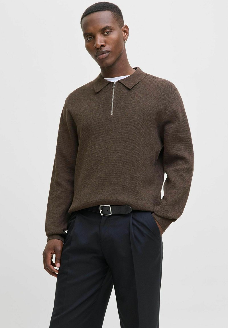 Muž v hnědém svetr se zipem, černém opasku a tmavých kalhotách, stojí s jednou rukou v kapse na prostém světlém pozadí.