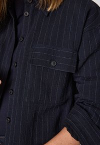 Camicia blu navy con bottoni, a righe verticali bianche, realizzata in tessuto testurizzato. Presenta una tasca sul petto e bottoni neri a contrasto.