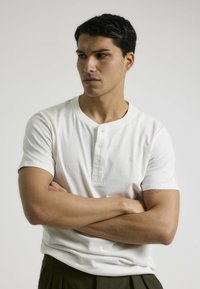 Camisa henley blanca de manga corta con tapeta de tres botones. Fabricada en un tejido suave. Presenta mangas ajustadas y un diseño de cuello redondo.