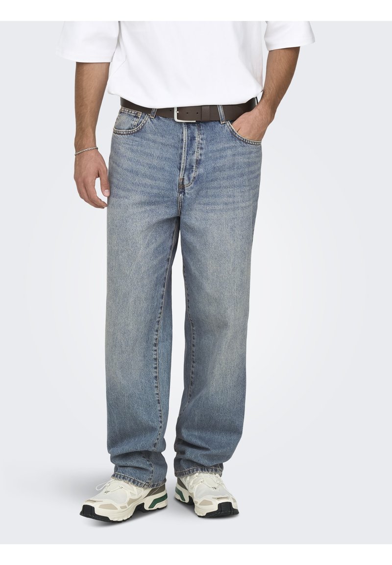 Jean en denim bleu à coupe décontractée, doté d'une braguette éclair, de cinq poches et d'un léger délavage. Porté avec une chemise blanche et des baskets beiges.