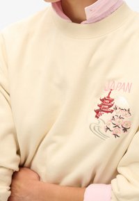 Felpa crema con colletto a coste, caratterizzata da una pagoda rossa ricamata, fiori di ciliegio e la scritta "GIAPPONE" in rosa. Tessuto morbido, vestibilità rilassata.