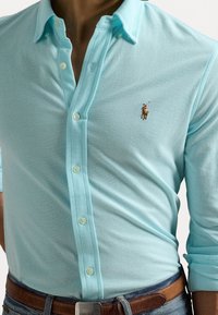 Muž v světle modré košili na knoflíky s vyšitým pólistou, hnědým páskem a vyhrnutými rukávy.