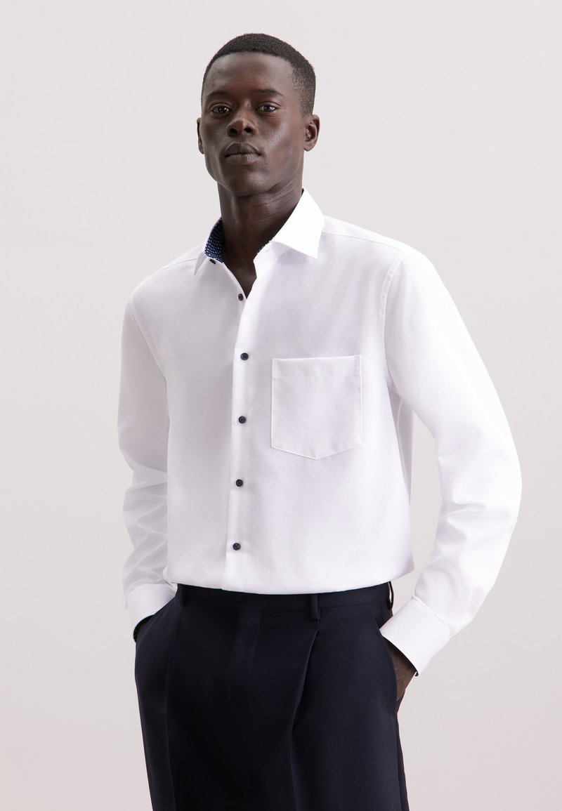Witte button-up shirt met een zak, zwarte knopen en een contrasterende binnenkraag met marineblauw patroon, gecombineerd met donkere broeken.