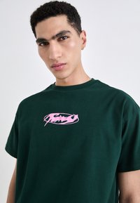 Grønn bomulls t-skjorte med rund hals og korte ermer. Har et rosa grafisk logo trykt i midten.