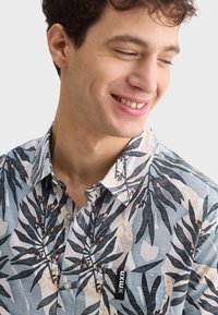 Košeľa s krátkym rukávom, zapínaná na gombíky v svetlomodrej farbe s tropickým vzorom listov v tmavozelenej a broskyňovej farbe, ktorá obsahuje náprsné vrecko s logom.