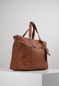 Bolso tote de piel marrón con dos asas superiores, un bolsillo frontal con cremallera y un acabado texturizado. Presenta un detalle de fleco para un estilo adicional.