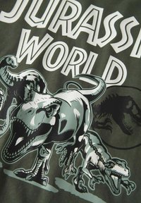 T-shirt en coton vert avec des graphiques de dinosaures en noir et blanc et le texte "Jurassic World", comprenant des illustrations détaillées du T. rex et d'autres.