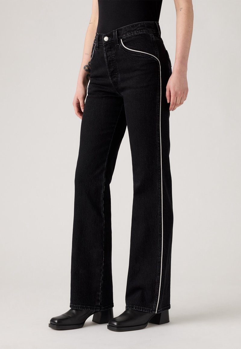 Personne portant un jean noir taille haute avec un liseré blanc sur le côté, associé à des bottes à talons noires et un haut noir ajusté.